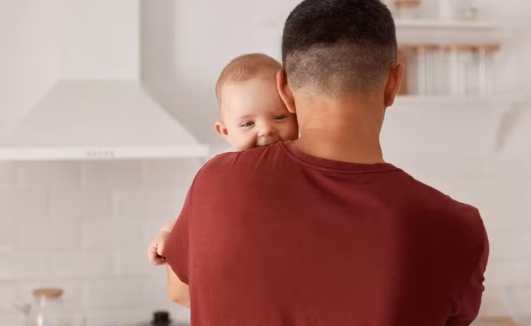 back-view-portrait-young-adult-caucasian-father-holding-son-daughter-cute-baby-looking-camera-mans-shoulder-posing-indoor-with-kitchen-set-background